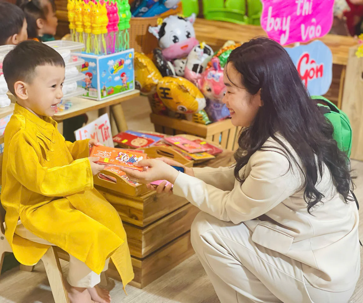 Ly kneeling to hand a red lucky envelope to a little boy in a yellow ao dai at a Tet fair